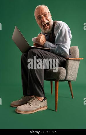 Full length portrait of senior man with beard in glasses, wearing vintage suit sitting on armchair with laptop looks happy and satisfied. Win. Stock Photo