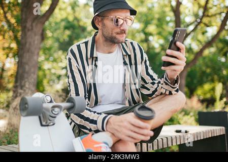 Adult bearded stylish guy relaxing in summer park after skateboarding, using internet on phone and drinking coffee Stock Photo
