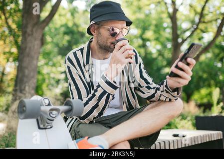 Portrait of adult male who drinking coffee and using phone at extreme park near skateboard, summer vibe. resting Stock Photo