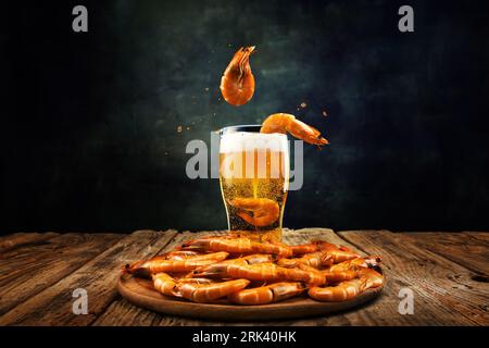 Delicious shrimp falling down into fresh, chill, foamy lager beer glass on wooden table over dark background Stock Photo