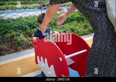 A cute two year old baby boy has fun playing on a colorful swing in the park, while his mom watches over his safety. Stock Photo