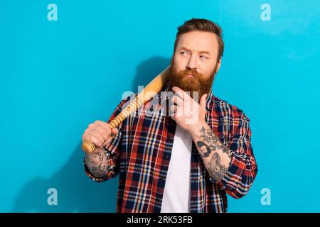 Photo of minded suspicious man hand on beard dressed checkered shirt look empty space bat on shoulder isolated on blue color background Stock Photo