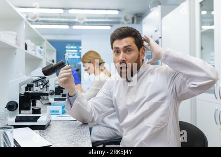 Portrait of a shocked young male scientist sitting in a laboratory behind a microscope, doing research and making a discovery. He holds the substance in his hand and is surprised by the result. Stock Photo