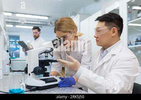 A diverse team of doctors and scientists works in the laboratory. Asian man and woman examine samples under microscope, study, discuss. Stock Photo