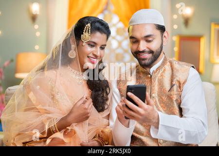 Happy smiling indian Muslim couple busy using mobile phone during Ramadan festival sales at home - concept of social media sharing, technology and Stock Photo