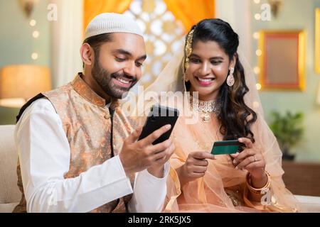 Indian happy smiling muslim couple making online payment for shopping using credit card on mobile phone during ramadan festival sales at home - Stock Photo