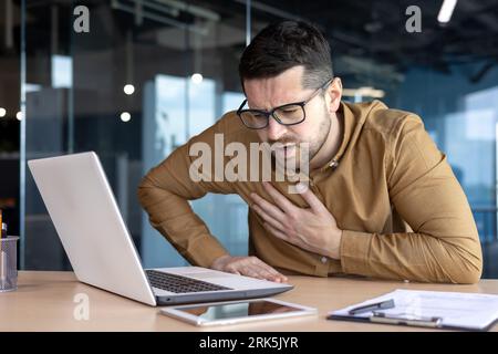 Heart attack, panic attack at work. A young man holds his chest while sitting at the desk in the office. Winced from pain, feels discomfort. Stock Photo