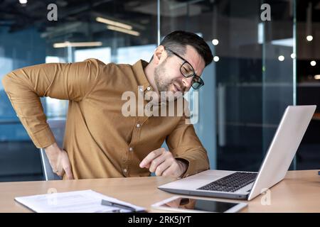 Injury at the workplace. A young man works in the office at a table behind a laptop and holds his hand behind his back. Feels severe pain, sprains, massages. Stock Photo