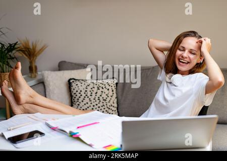 Young laughing female student in white t-shirt studying at home, using laptop e-learning and having fun. Stock Photo