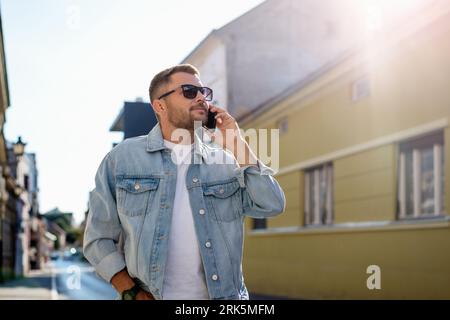 Handsome young urban man in trendy denim jacket and sunglasses talking on the phone while walking street of European town. Stock Photo