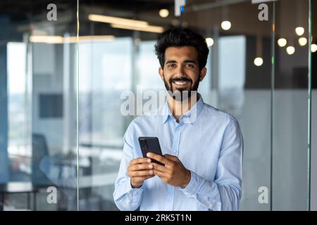 Portrait of Indian young male student standing inside campus, office, university and using phone. Smiling and looking at the camera. Stock Photo
