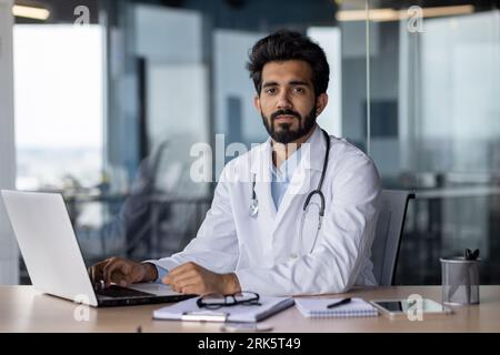 Portrait of a young doctor student studying online at a laptop, sitting in the office in a white coat. He looks seriously into the camera. Stock Photo