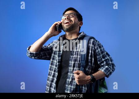 Cheerful young adult college student wearing a backpack and talking on a mobile phone against a vivid blue sky background Stock Photo