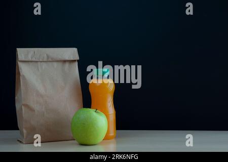 paper bag with the snack for school with a green apple and a bottle of orange juice on the desk on a black background of the chalkboard with a place f Stock Photo