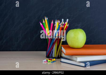 metal holder with crayons and green apple and books on desk on black  chalkboard background Stock Photo