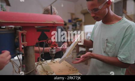 Experienced master carpenter guiding young apprentice at carpentry workshop. Brazilian student giving thumbs up to boss nexto to industrial machine Stock Photo