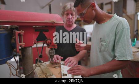 Experienced master carpenter guiding young apprentice at carpentry workshop. Brazilian student giving thumbs up to boss nexto to industrial machine Stock Photo