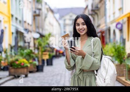 Beautiful young Indian woman using phone and credit card. Standing outside on a city street and smiling at the camera. Stock Photo