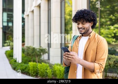Young smiling Indian male student, freelancer, tourist standing outside and holding phone. Writes a message, looks for a way, calls. Stock Photo