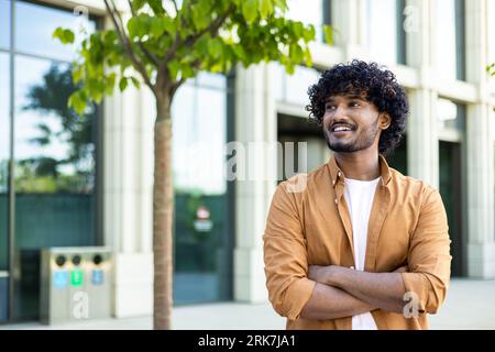 Portrait of a successful young male designer, engineer standing outside an office center, crossing his arms on his chest and looking to the side with a smile. Stock Photo