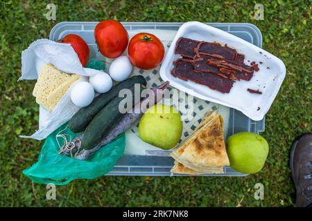 Food for the hiking group lies on the lid of a plastic box in the grass in the morning in Dusheti Municipality, Mtskheta-Mtianeti region, Georgia. A piece of cheese and three eggs lie on a paper towel. Next to them lie three tomatoes, two cucumbers and a purplish-brown stick on a string (possibly churchkhela). Slices of dried meat lie in a polystyrene tray. Two green apples and folded flatbread lie beside them. The tour was organised by the agency Enjoy Georgia, with Giorgi Chachua as hiking guide. The trekking tour with pack horses covered almost 120 kilometres and more than 5,000 metres of a Stock Photo