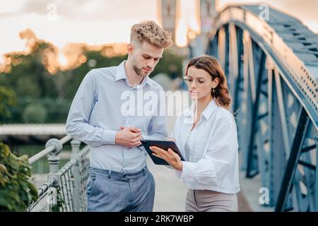 Business professionals are standing on a bridge together at sunset, intently discussing something on a tablet Stock Photo