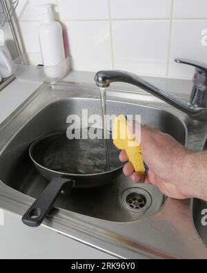 Washes the frying pan in the kitchen under running water. Man's hand with yellow washing sponge Stock Photo