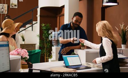 Diverse couple checking in at hotel, talking to concierge about reservation and receiving access key card at reception. Front desk staff helping guests, providing luxury service. Handheld shot. Stock Photo
