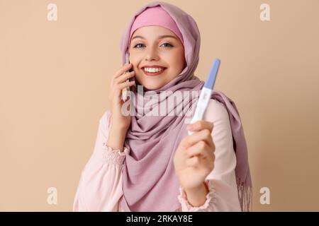 Happy Muslim young woman with positive pregnancy test talking by phone on beige background Stock Photo