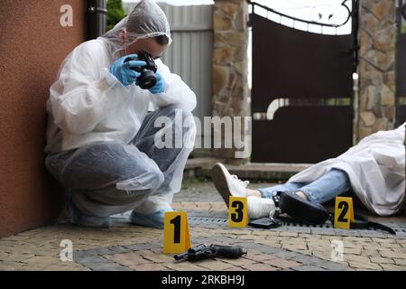 Criminologist taking photo of evidence at crime scene with dead body near house Stock Photo