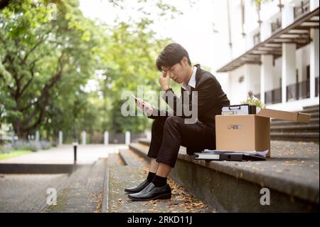 A stressed and upset young Asian businessman sits on a staircase with a box of his personal stuff after getting fired or losing his job. Unemployed, c Stock Photo