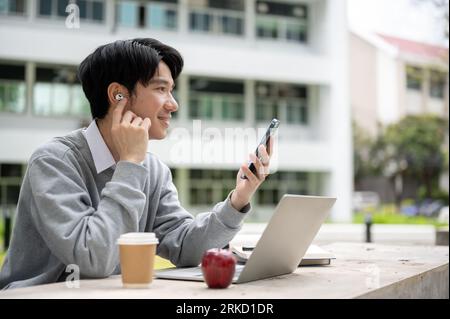 A happy and handsome young Asian male college university student enjoys listening to music through his earbuds while doing homework at a table in a ca Stock Photo
