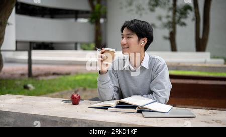 A happy and relaxed young Asian male college student enjoys his coffee and listens to music while preparing for an exam in a campus park. Stock Photo