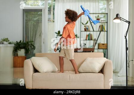 Boy standing on couch and playing with toy plane imagining himself as a pilot Stock Photo