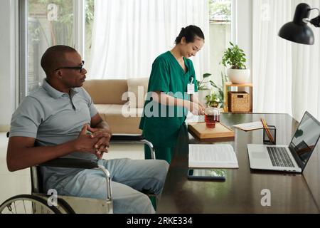 Social worker making tea for man with disability sitting in wheelchair Stock Photo