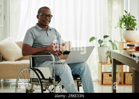 Portrait of smiling Black man with laptop sitting in wheelchair in his apartment Stock Photo