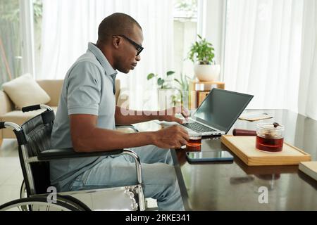 Black man with disability sitting at desk, drinking tea and checking e-mails on laptop Stock Photo