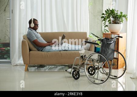 Man with disability sitting on couch at home and working or playing on laptop Stock Photo