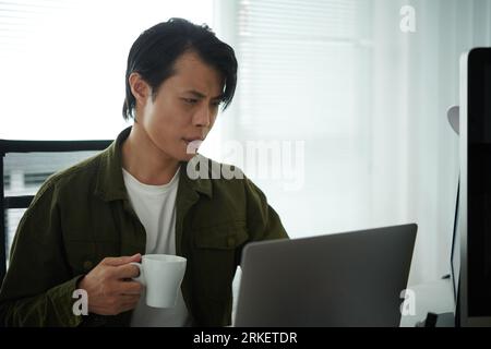Frowning software developer drinking morning coffee and checking e-mails from clients and coworkers on laptop Stock Photo