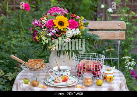 table arrangement with cup of tea, apple cake, apples in basket and bouquet of summer flowers Stock Photo