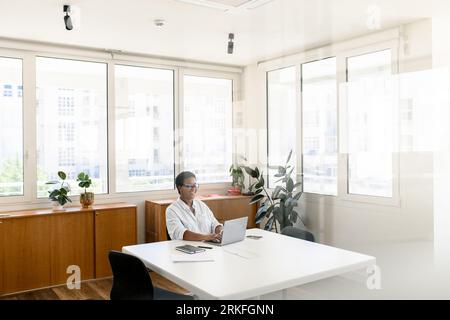 Cheerful African-American female office employee using laptop sitting in the modern coworking space, woman in casual wear and glasses typing indoor, manager on the workplace Stock Photo