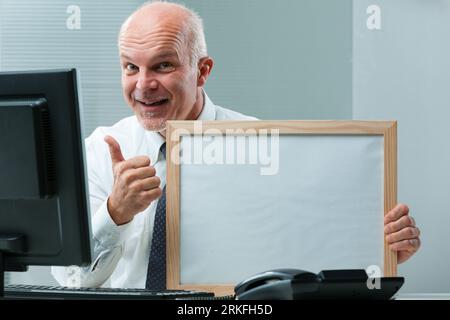 enthusiastic senior businessman at a desk holds an empty sign with COPYSPACE for your content. His reassuring smile and wink suggest a win-win offer b Stock Photo