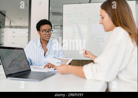 Two businesswomen sitting at the desk and discussing. Diverse female office employees brainstorming in meeting room. HR manager interviewing candidate for the position Stock Photo