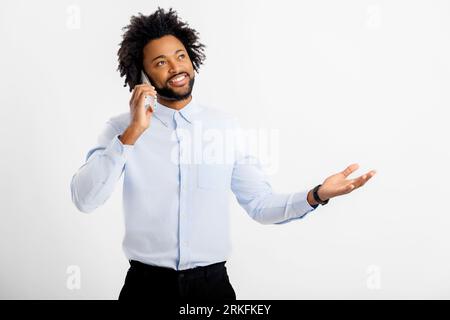 Always in touch. African-American curly man in white formal shirt talking on the smartphone and gesturing, having conversations with customers or colleagues, studio shot isolated on white Stock Photo