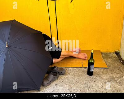 Poor and drunk man sleeping on behind the umbrella at sidewalk with bottles of wine near Stock Photo