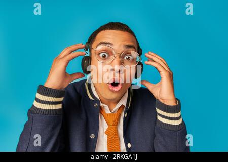 impressed african american student in eyeglasses and headphones, with open mouth on blue Stock Photo