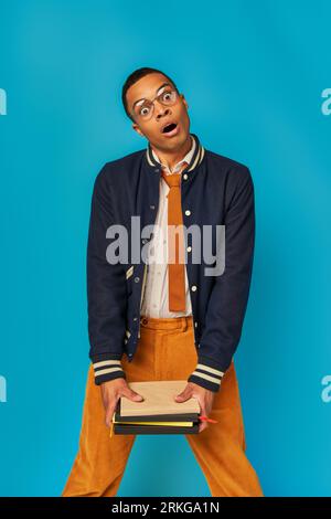 shocked african american student in stylish clothes holding notebooks and looking at camera on blue Stock Photo