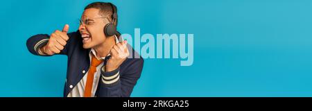 thrilled african american student in wireless headphones showing thumbs up on blue, banner Stock Photo