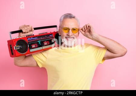 Photo of toothy beaming man with beard dressed yellow t-shirt holding boombox on shoulder touch sunglass isolated on pink color background Stock Photo