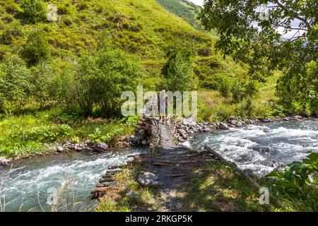Three hikers cross a walkway of tree trunks and stones over a mountain river in single file near the village of Ardoti in Dusheti Municipality, Mtskheta-Mtianeti region, Georgia. Foaming water rushes past on both sides of the walkway. Bushes and trees grow on the bank. A green slope rises behind them. The trekking tour with pack horses covered almost 120 kilometres and more than 5,000 metres of ascent. The largely abandoned village lies on a hill above the Andaki valley, around 5 kilometres south-west of Mutso. Khevsureti (Georgian ხევსურეთი) is a historical region on the slopes of the Greater Stock Photo